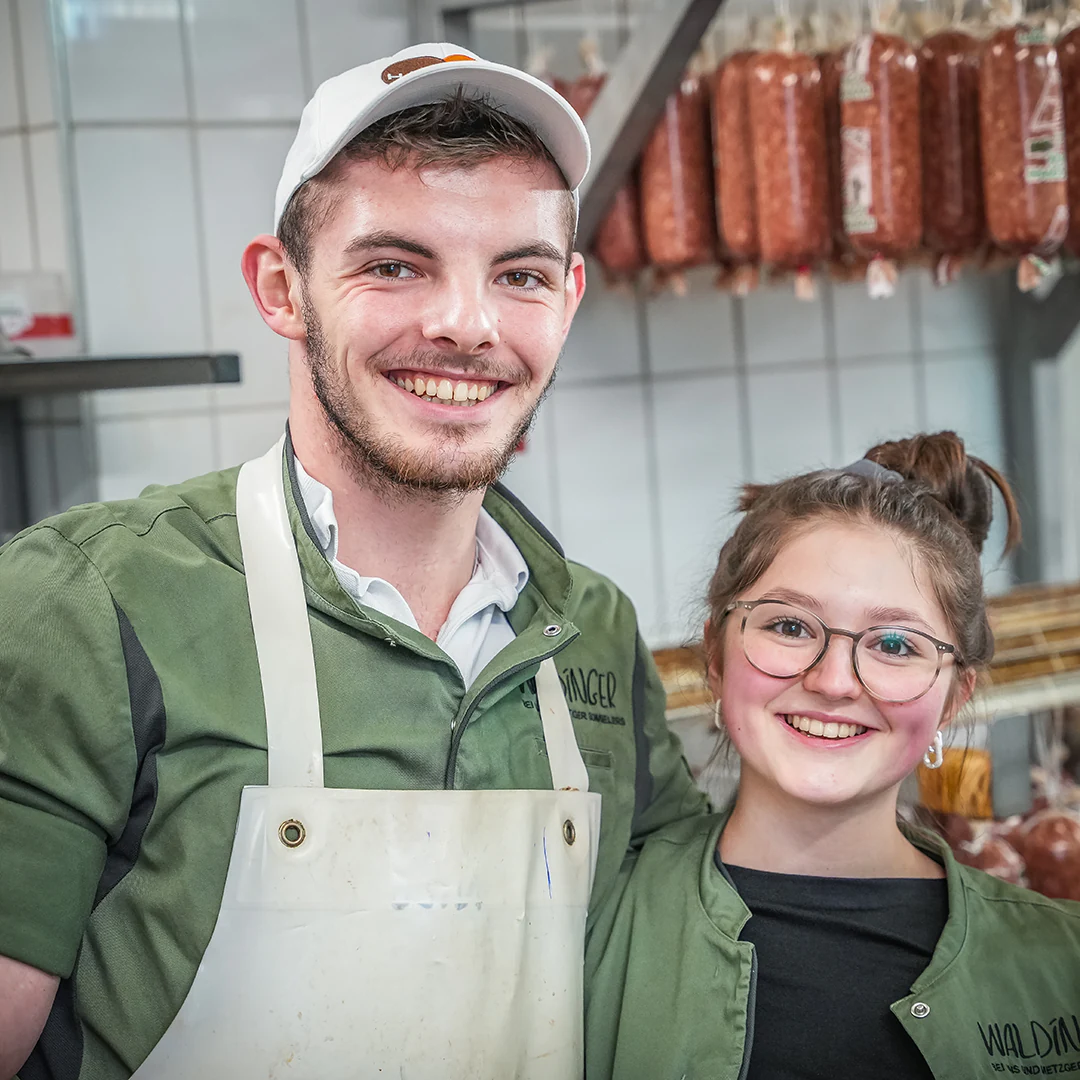 Zwei junge Metzger in grüner Waldinger-Arbeitskleidung lächeln in einer Wurstkammer, umgeben von hängenden Wurstwaren.