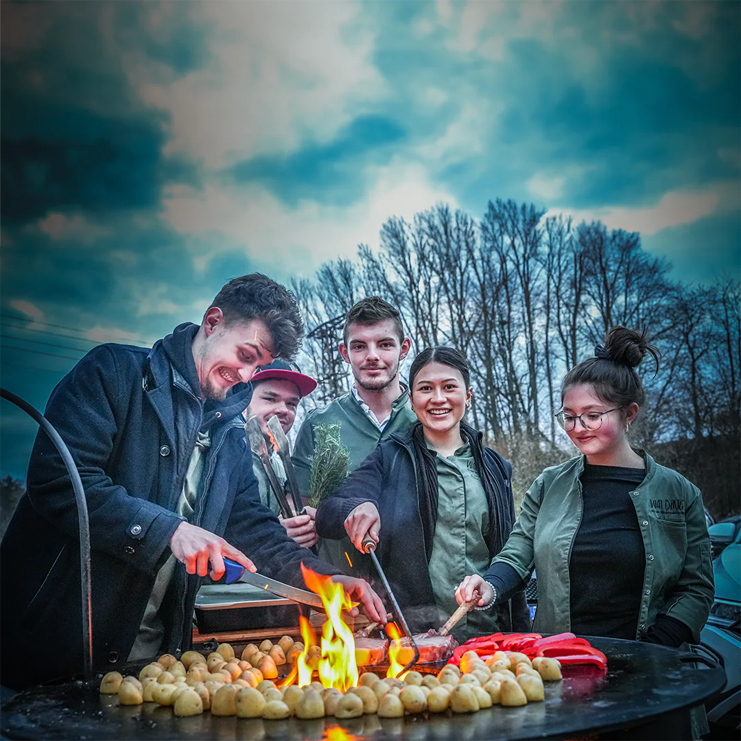 Ein Team von jungen Metzgern grillt gemeinsam im Freien, mit Fleisch, Kartoffeln und Gemüse auf einer Feuerplatte.