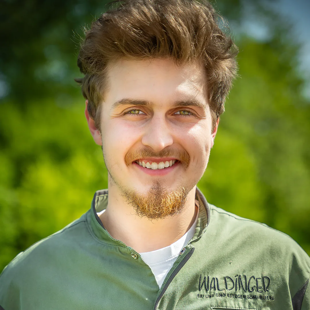 Porträt eines jungen Mannes mit grünem Metzgerkittel von Waldinger, lächelnd vor einem natürlichen, grünen Hintergrund.