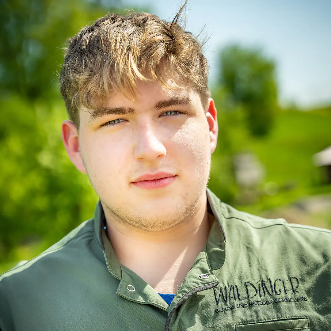 Porträt eines jungen Mannes mit kurzem, blondem Haar und grünem Waldinger-Kittel, ernst blickend vor ländlicher Kulisse.