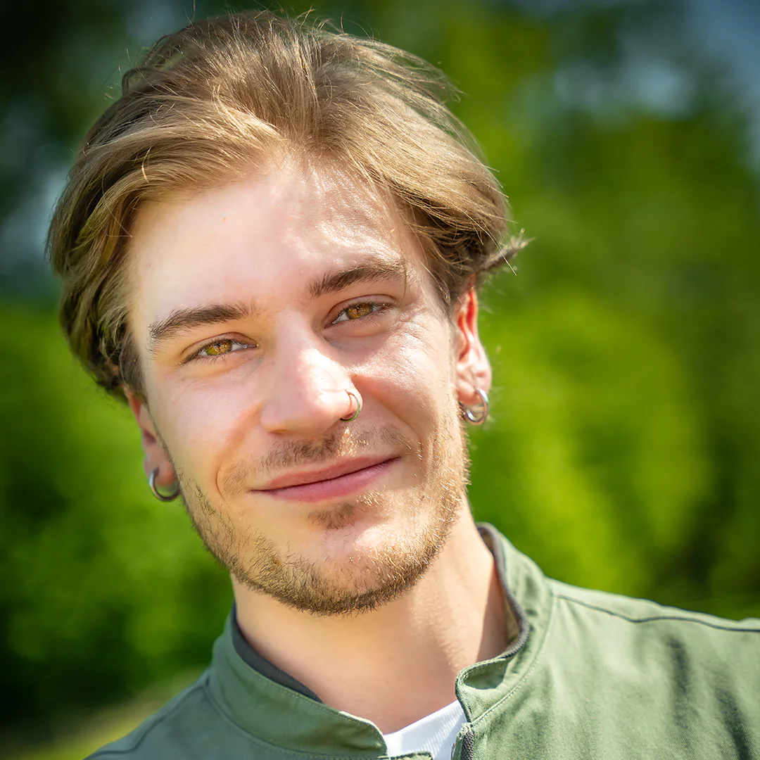 Porträt eines Mannes mit Ohrringen und grünem Waldinger-Kittel, mit leichtem Lächeln vor grüner Landschaft.