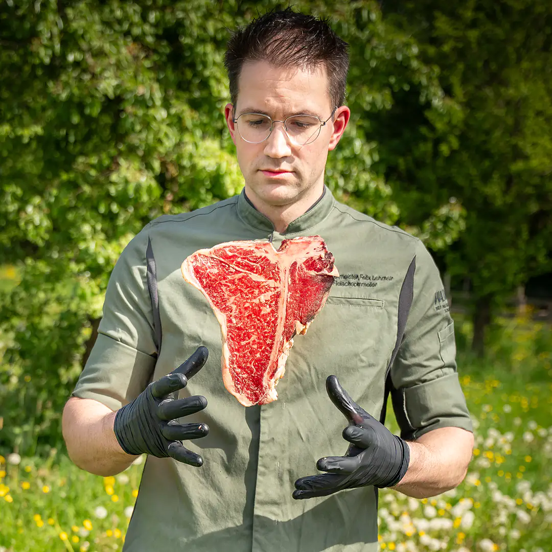 Metzger in grüner Waldinger-Arbeitskleidung und schwarzen Handschuhen wirft ein perfekt marmoriertes T-Bone-Steak in die Luft und ist im Begriff, es aufzufangen, vor einer grünen Naturkulisse.