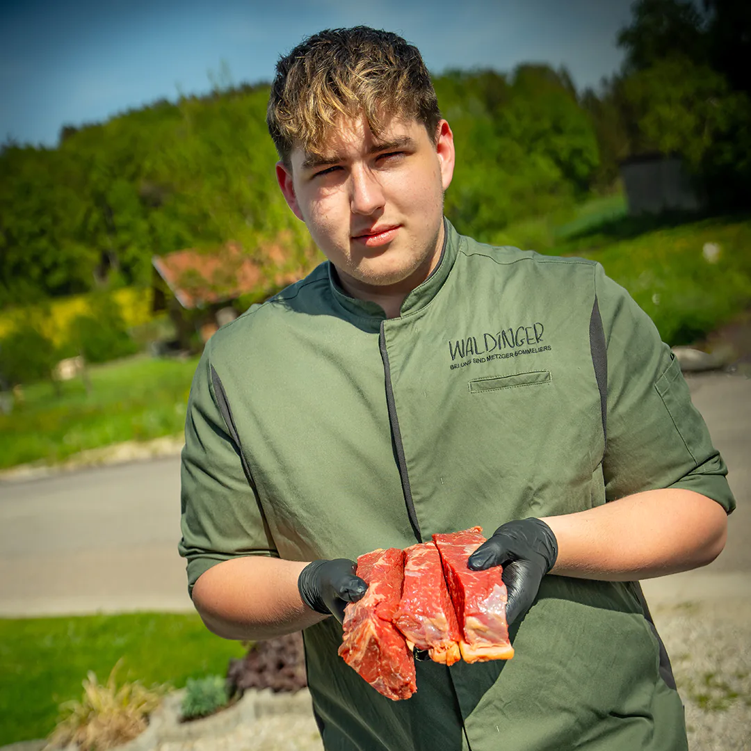 Ein junger Metzger in grüner Waldinger-Arbeitskleidung hält drei dick geschnittene Stücke Roastbeef in schwarzen Handschuhen, vor einer ländlichen Kulisse.