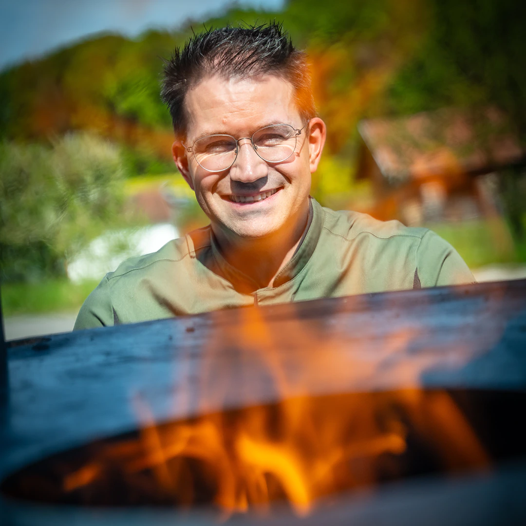 Metzger in grüner Arbeitskleidung lächelt hinter einer offenen Feuerstelle, während Flammen im Vordergrund lodern.