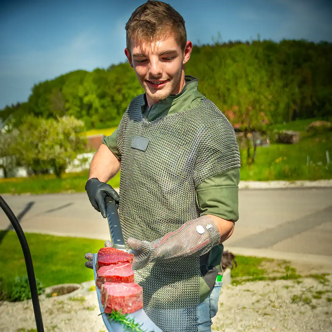 Metzger in Kettenhemd und Handschuhen balanciert mehrere Fleischstücke auf einer langen Messerklinge im Freien.