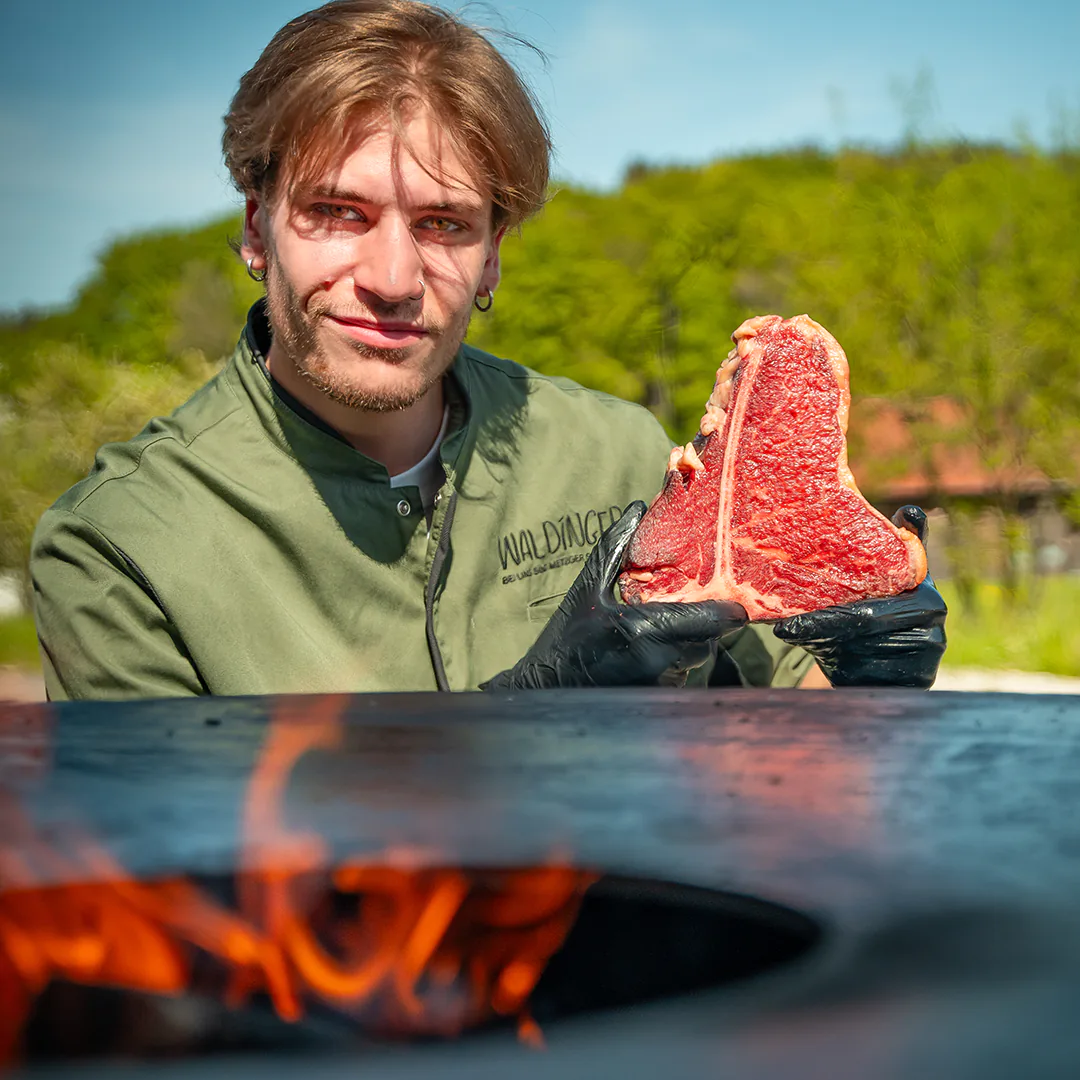 Metzger hält ein marmoriertes T-Bone-Steak mit schwarzen Handschuhen vor einer Feuerstelle, bereit zum Grillen.
