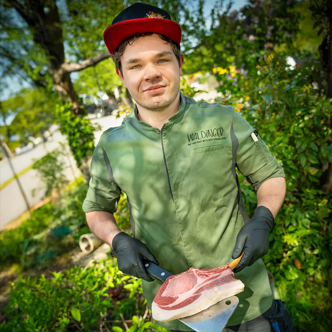Metzger mit roter Cap hält ein Tomahawk-Steak auf einem Beil und lächelt in die Kamera.