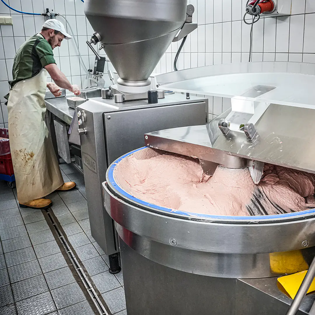 Wurstproduktion in der Metzgerei mit einer großen Kuttermaschine, die fein zerkleinertes Brät verarbeitet, während ein Mitarbeiter im Hintergrund arbeitet.