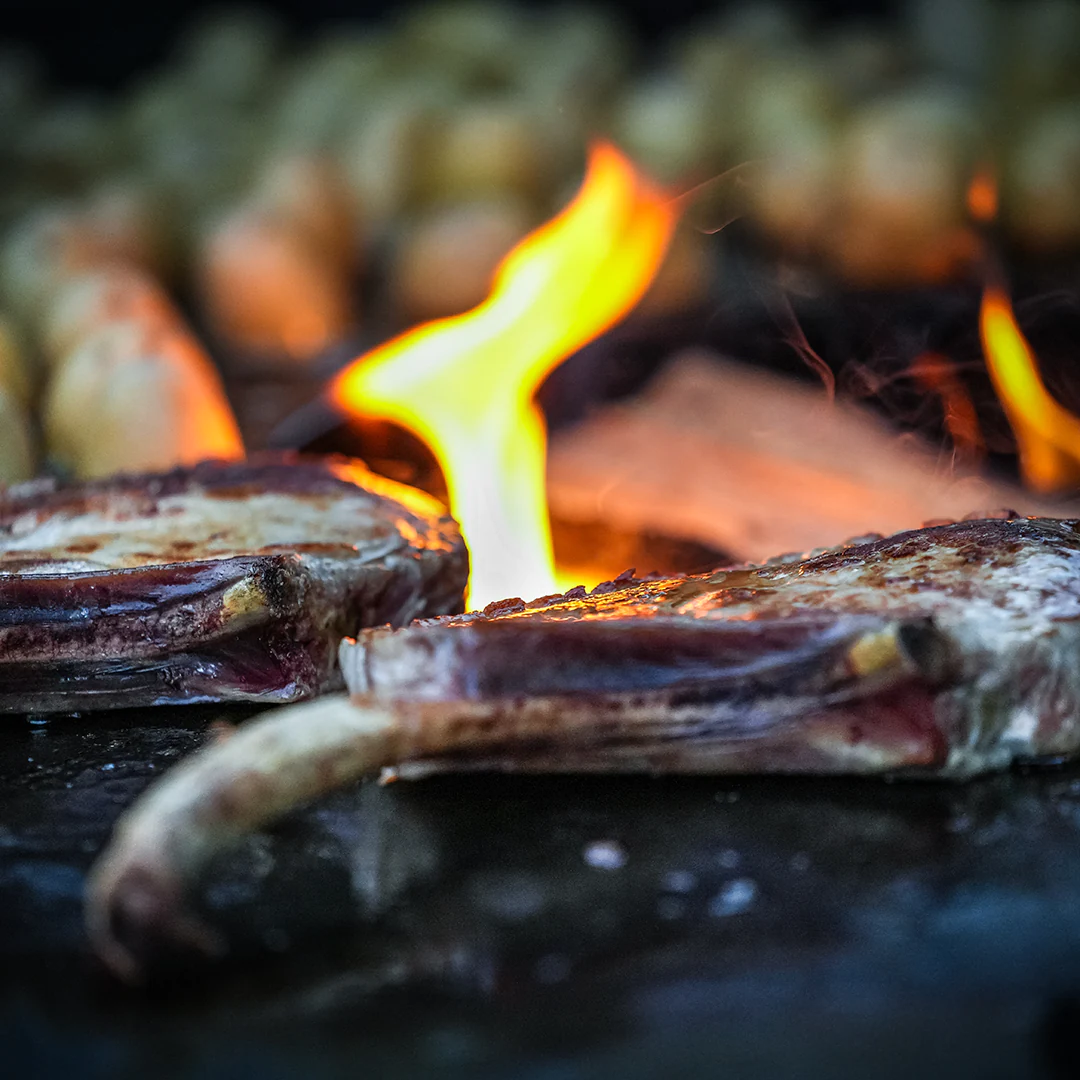 Zwei saftige Tomahawk-Steaks mit dunkler Kruste auf einem Grill, während im Hintergrund offene Flammen lodern und Kartoffeln rösten.