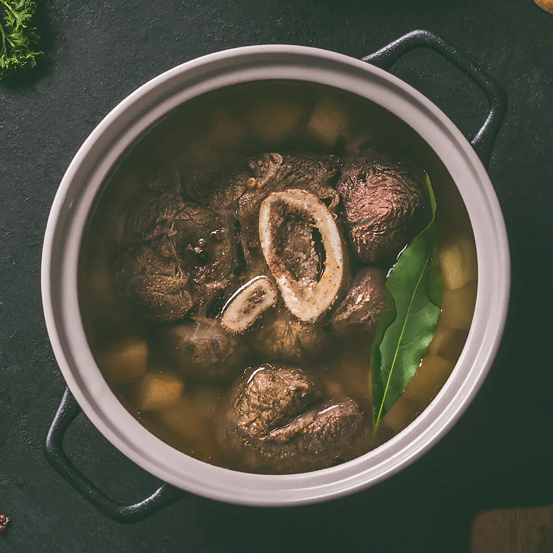 Topf mit kräftiger Fleischbrühe, durchzogen von Markknochen, Rindfleisch und Lorbeerblättern, auf dunkler Oberfläche angerichtet.