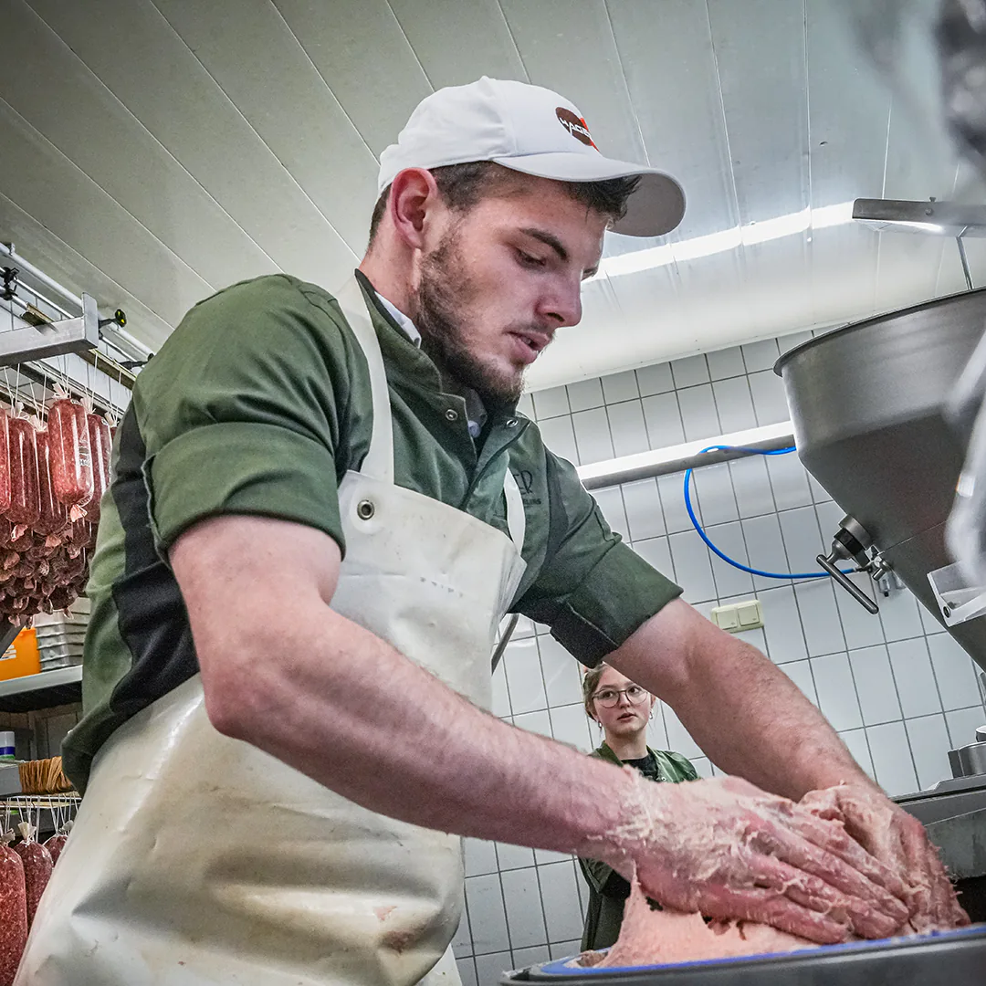 Ein Metzger in grüner Arbeitskleidung und Schürze knetet frisches Brät in der Wurstproduktion, während im Hintergrund Wurstwaren hängen und eine Kollegin zusieht.